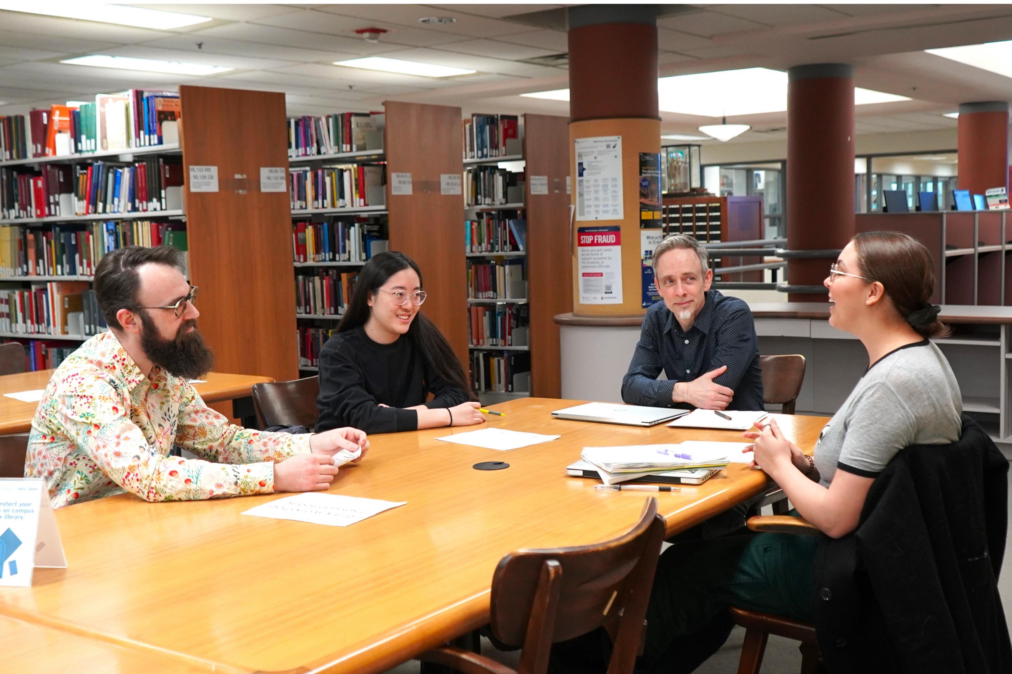 Two graduate students sit in the Music Library, animatedly discussing their research with Dr. Tim Neufeldt and Dr. Ely Lyonblum.